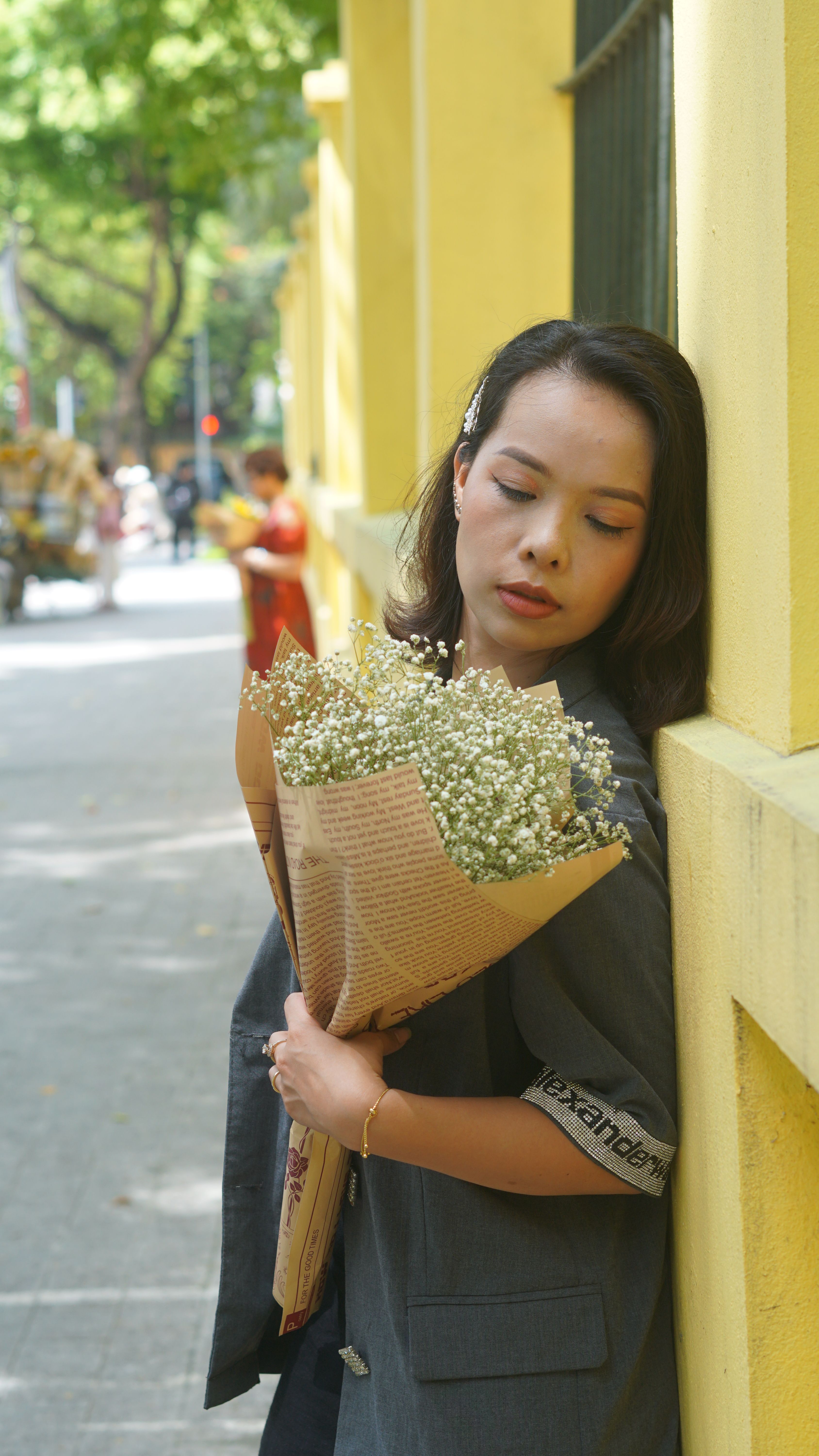 Woman holding flowers, standing against a vibrant yellow wall in an urban setting.