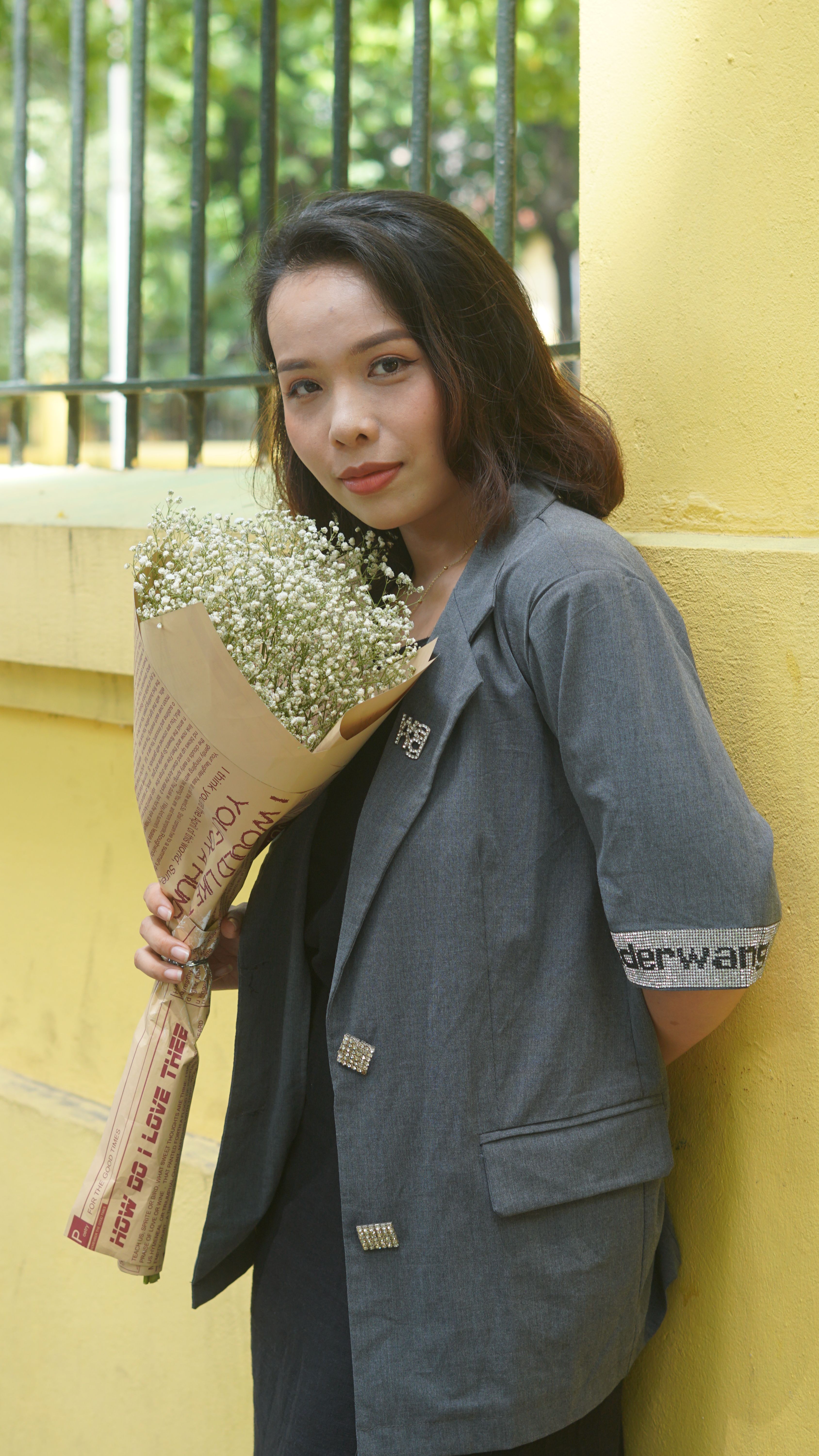 A young woman stands outdoors holding a delicate bouquet wrapped in newspaper.