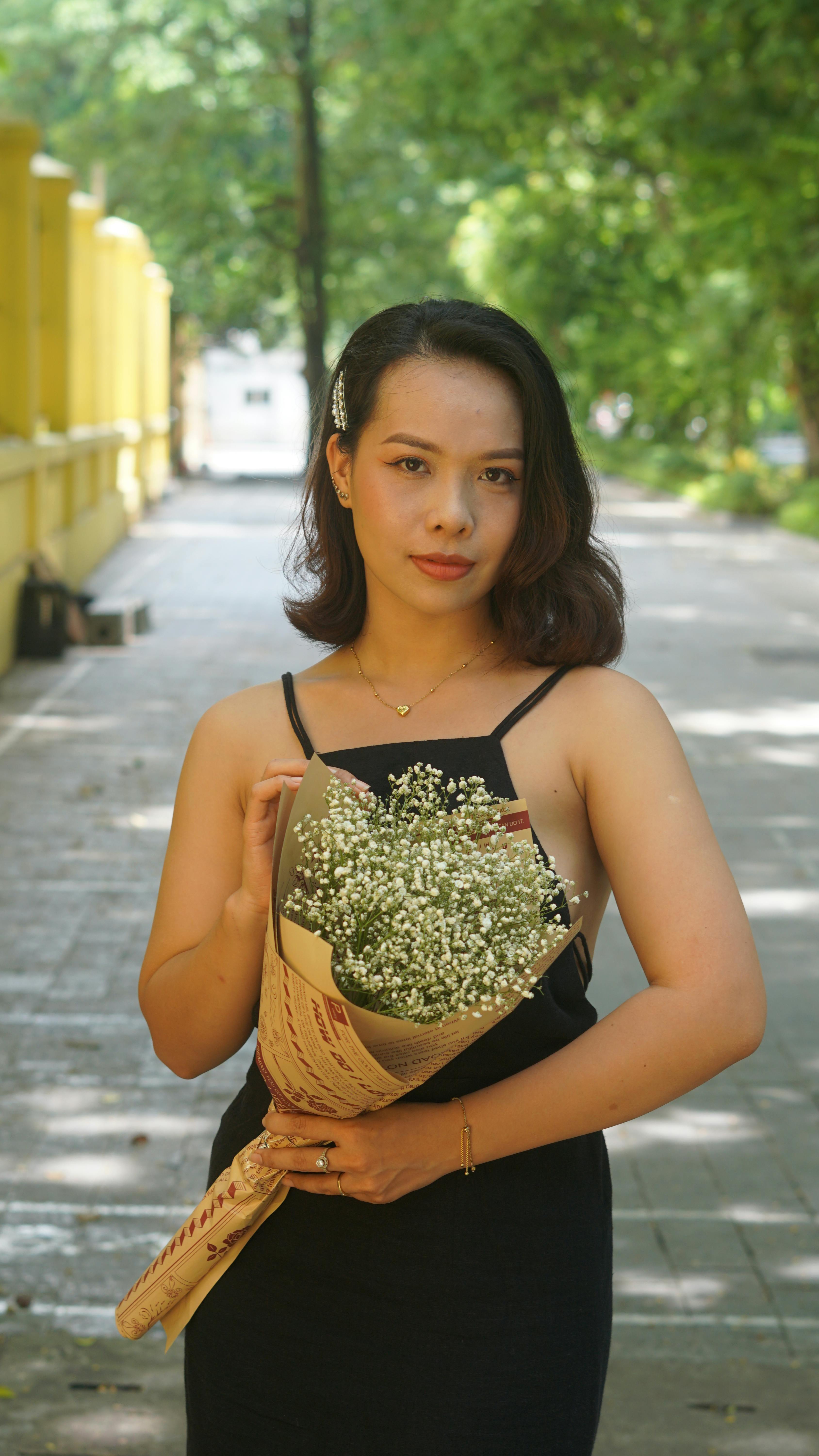 A stylish woman in a black dress holding a bouquet of flowers in a sunny outdoor setting.