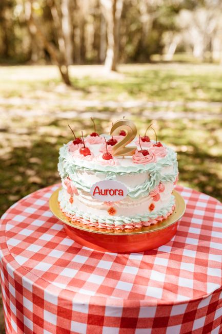 A vibrant birthday cake with cherries and flowers set on a gingham table outdoors.