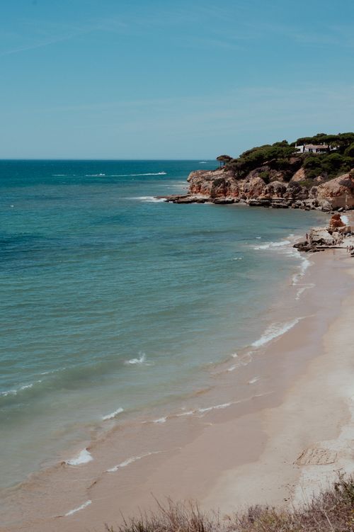 Free A picturesque coastal scene with sandy beach, gentle waves, and rocky cliffs under a clear blue sky. Stock Photo