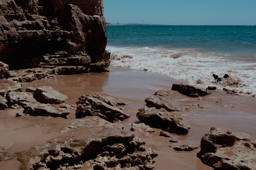 Free A scenic view of rocky cliffs and waves crashing on a sandy beach under a clear blue sky. Stock Photo