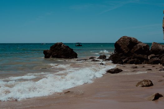 Discover the serene beauty of a rocky beach with waves gently crashing against sandy shores under a clear blue sky.
