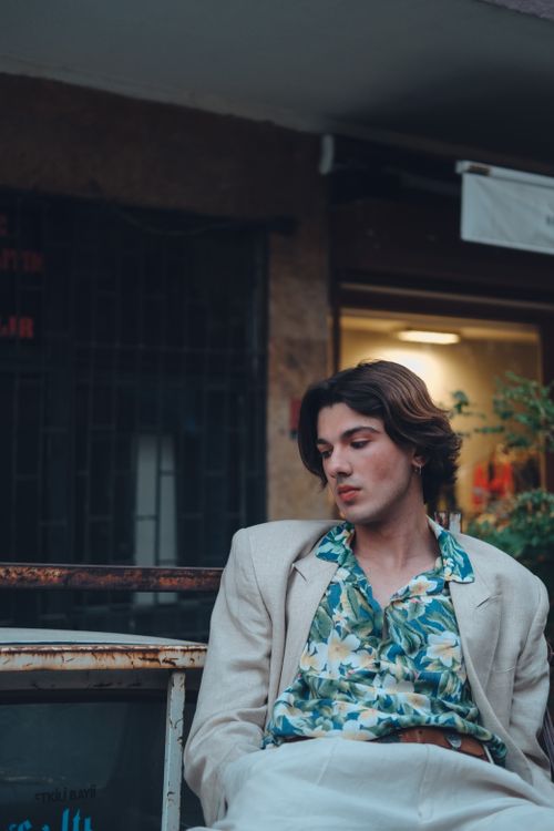 Free A fashionable man in a floral shirt and linen suit sits outdoors with a relaxed expression, combining elegance with a casual urban vibe. Stock Photo