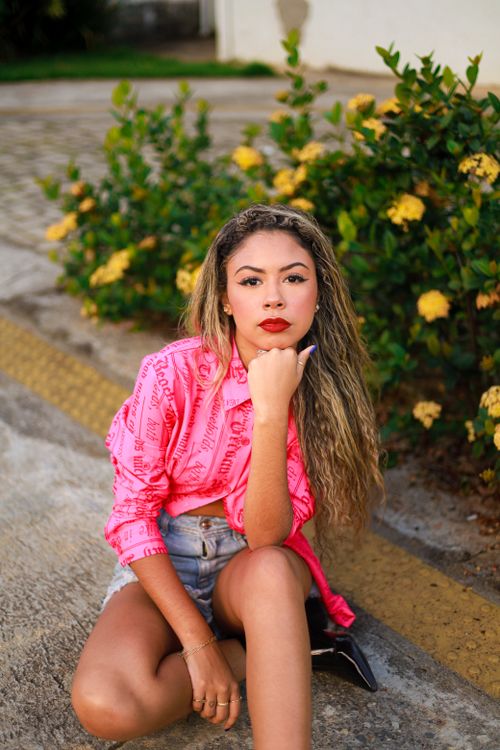 Free Stylish woman in a pink shirt and denim shorts sitting outdoors surrounded by yellow flowers. Stock Photo