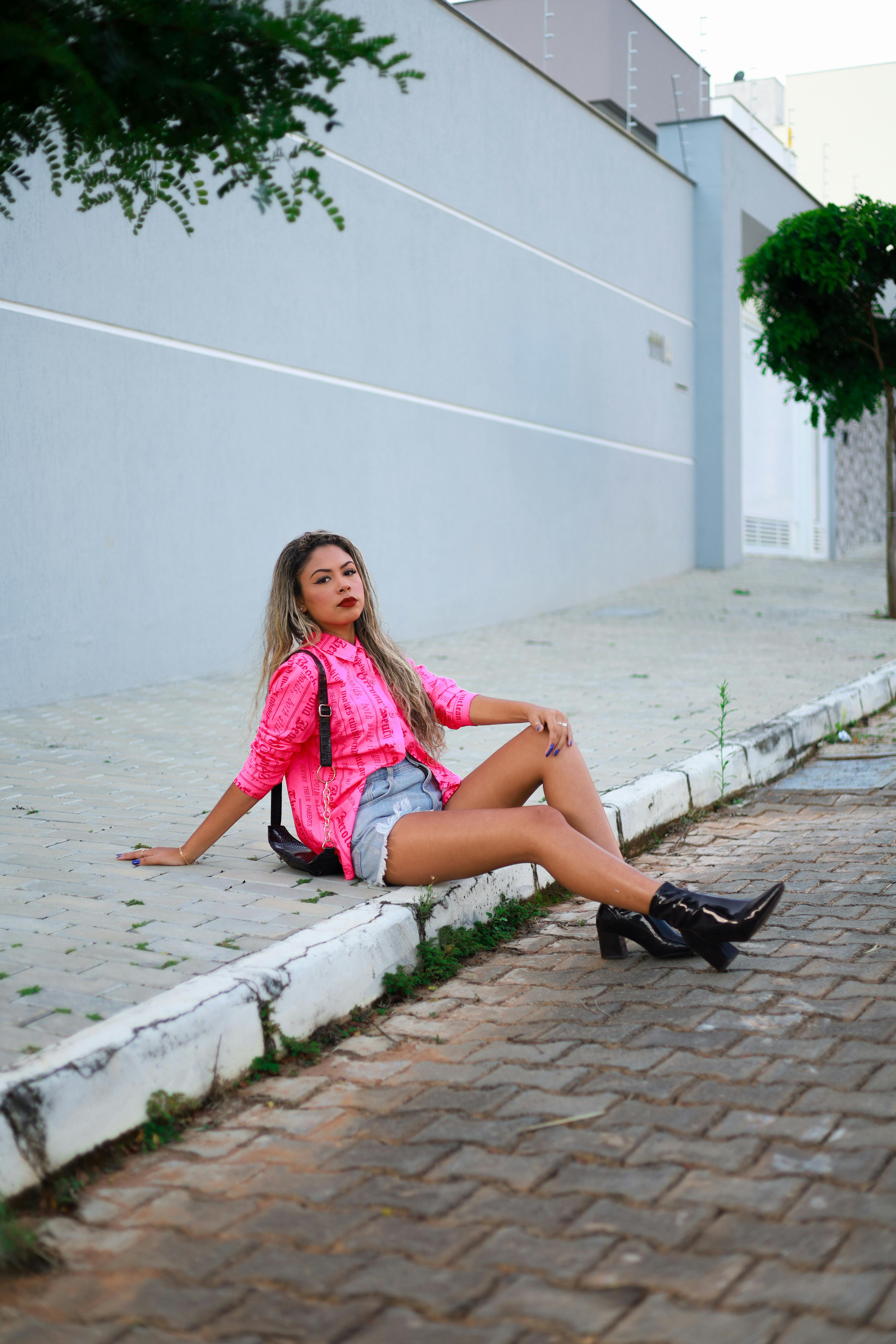 Fashionable woman in pink jacket and boots sitting on a city sidewalk, showcasing street style.