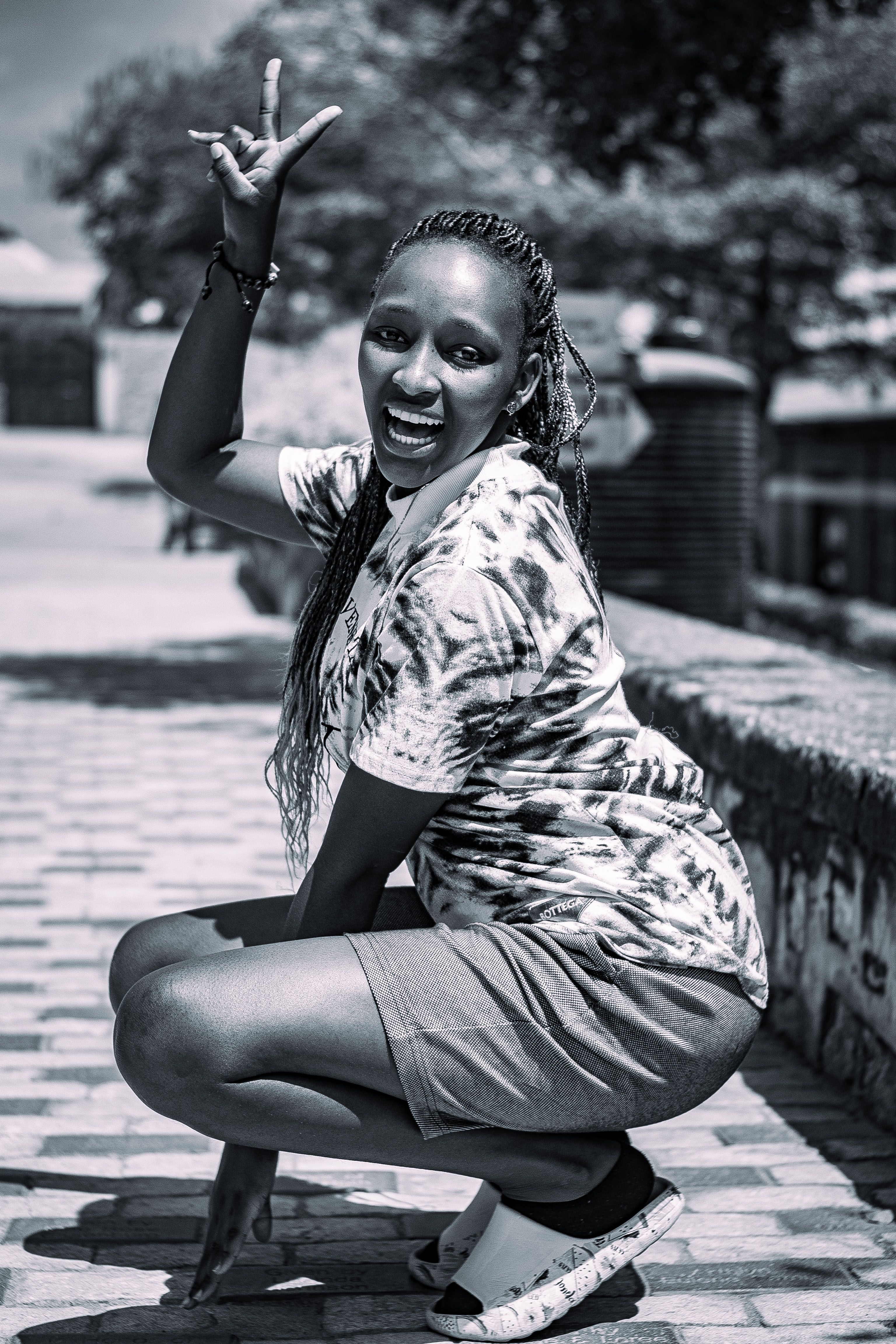 Black and white image of a smiling woman with braids, crouching and gesturing in an urban outdoor setting.