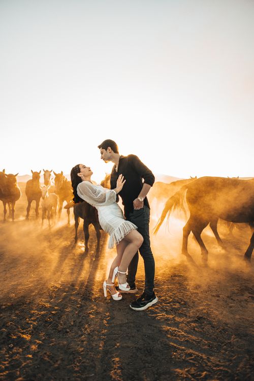 Free A couple shares a romantic moment amidst a herd of horses at sunset, creating a dreamy atmosphere. Stock Photo