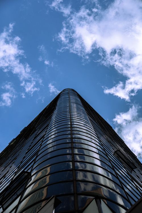Free Low-angle view of a modern glass skyscraper reflecting clouds and sky, symbolizing urban architecture. Stock Photo