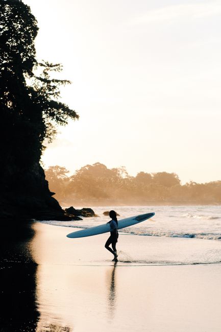 A silhouette of a woman carrying a surfboard along a tropical beach at sunset, reflecting tranquil vibes.