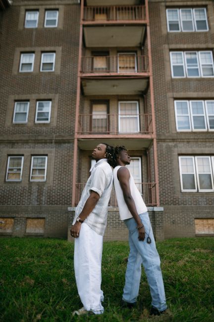 Two young men stand back to back in front of an apartment building, showcasing trendy fashion outdoors.