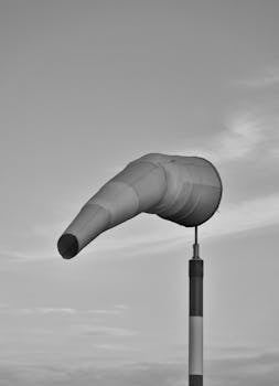 A black and white photo of a windsock blowing in the wind against a cloudy sky.