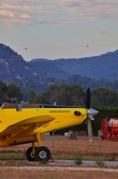 A vintage yellow propeller plane parked on a grassy airport runway with mountains in the background.