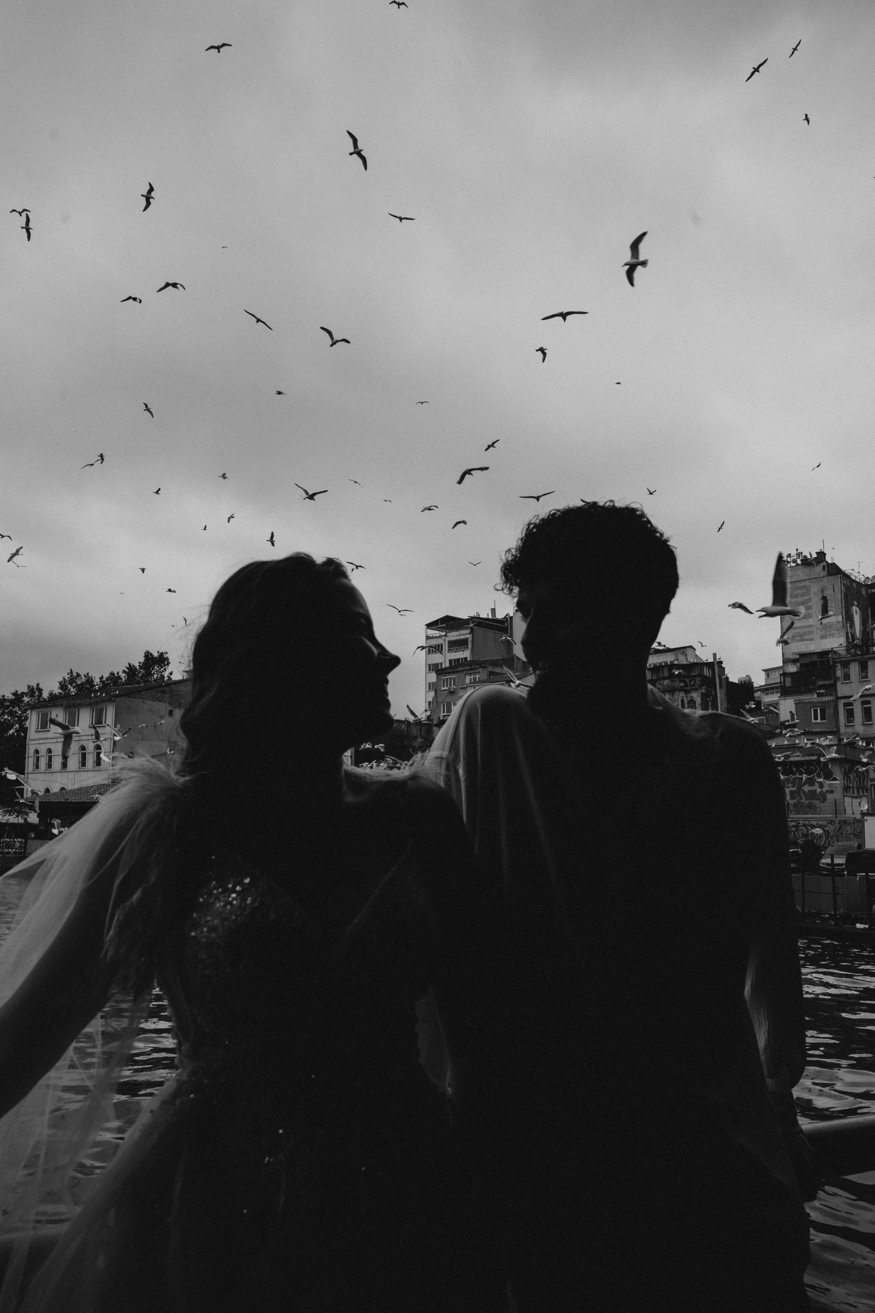 a couple standing in front of the water with birds flying around them