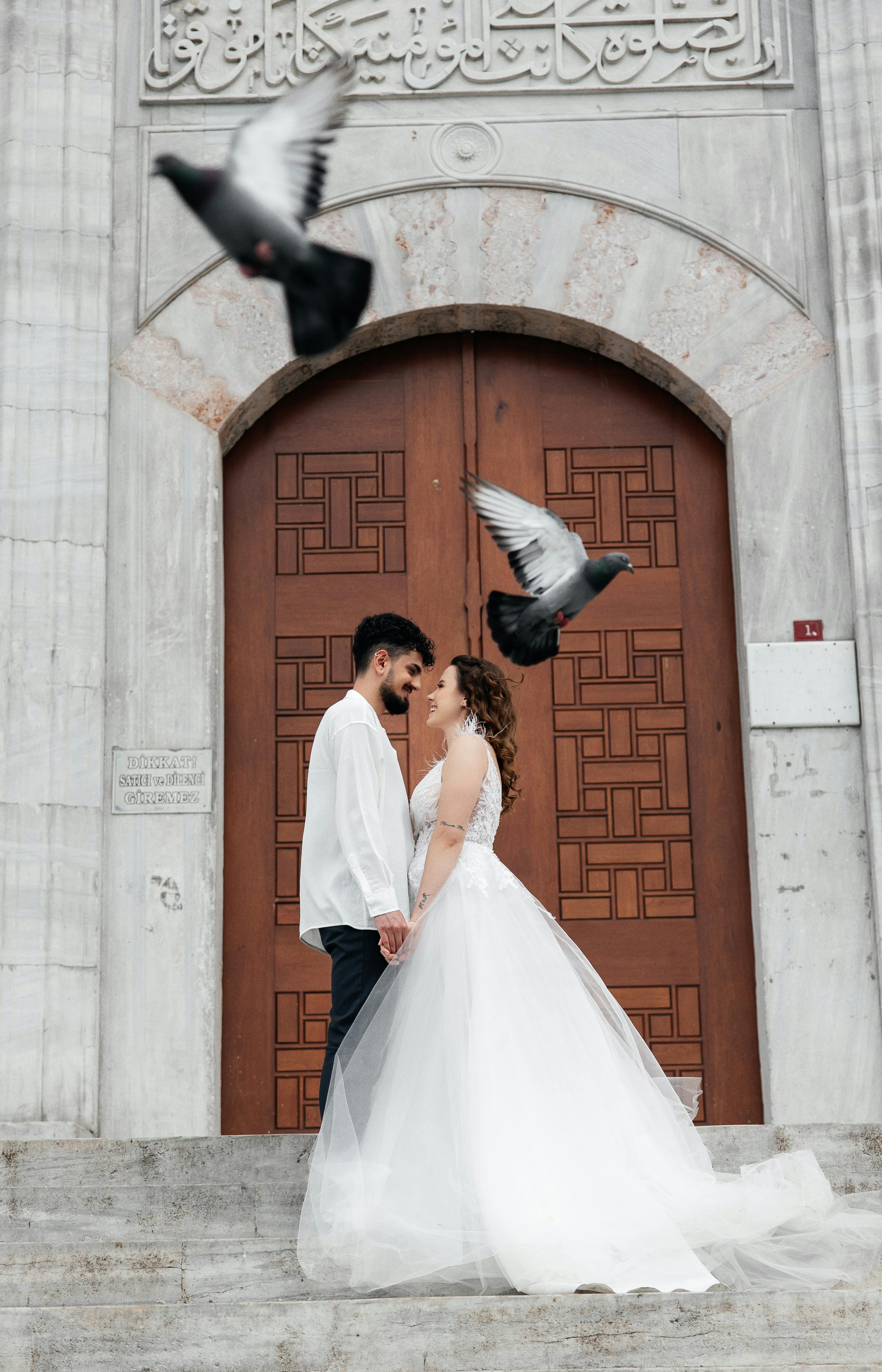 a bride and groom standing in front of a church with pigeons flying around them