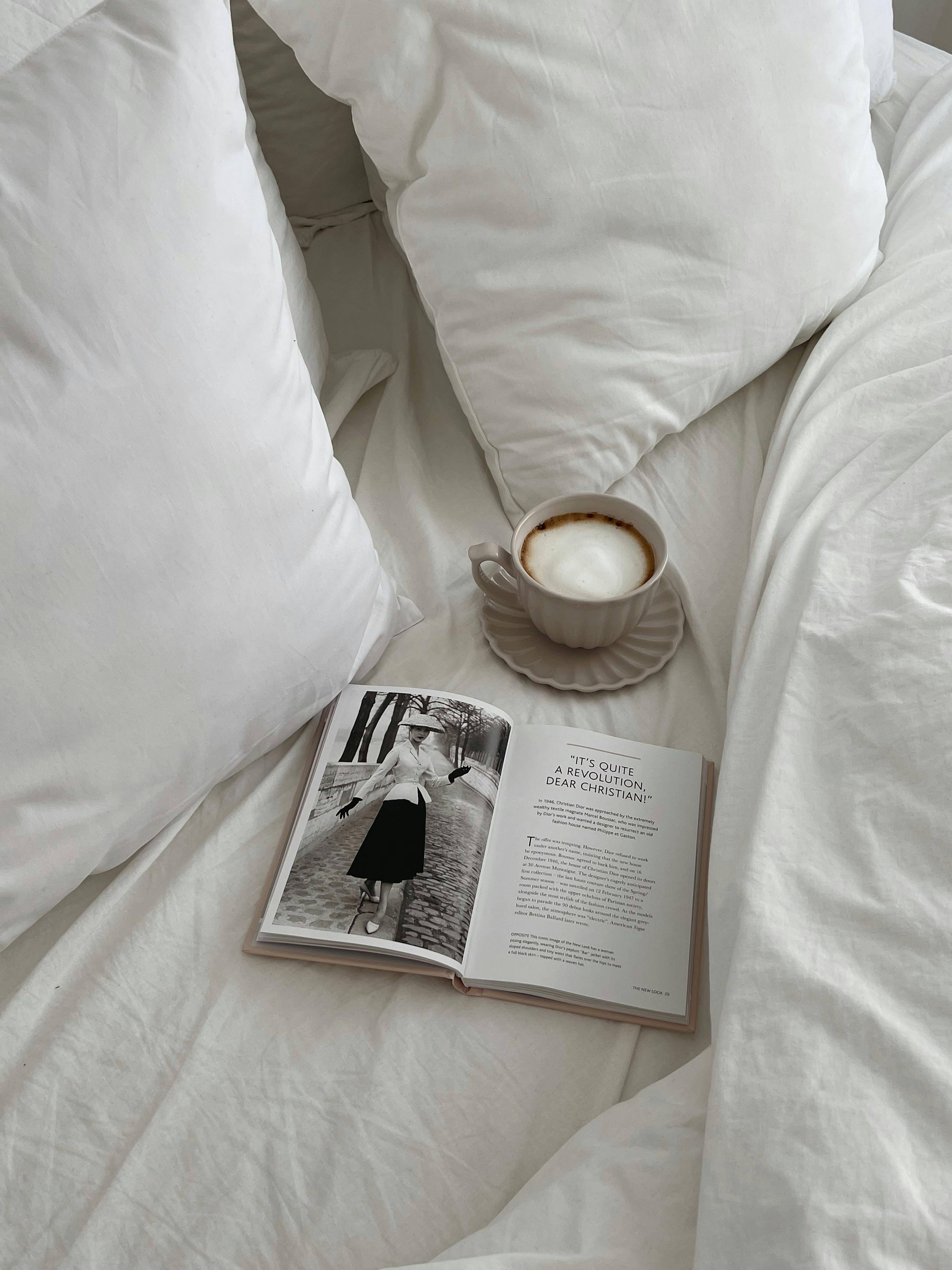Relaxing morning scene with a cappuccino and open book on bed with soft white pillows.
