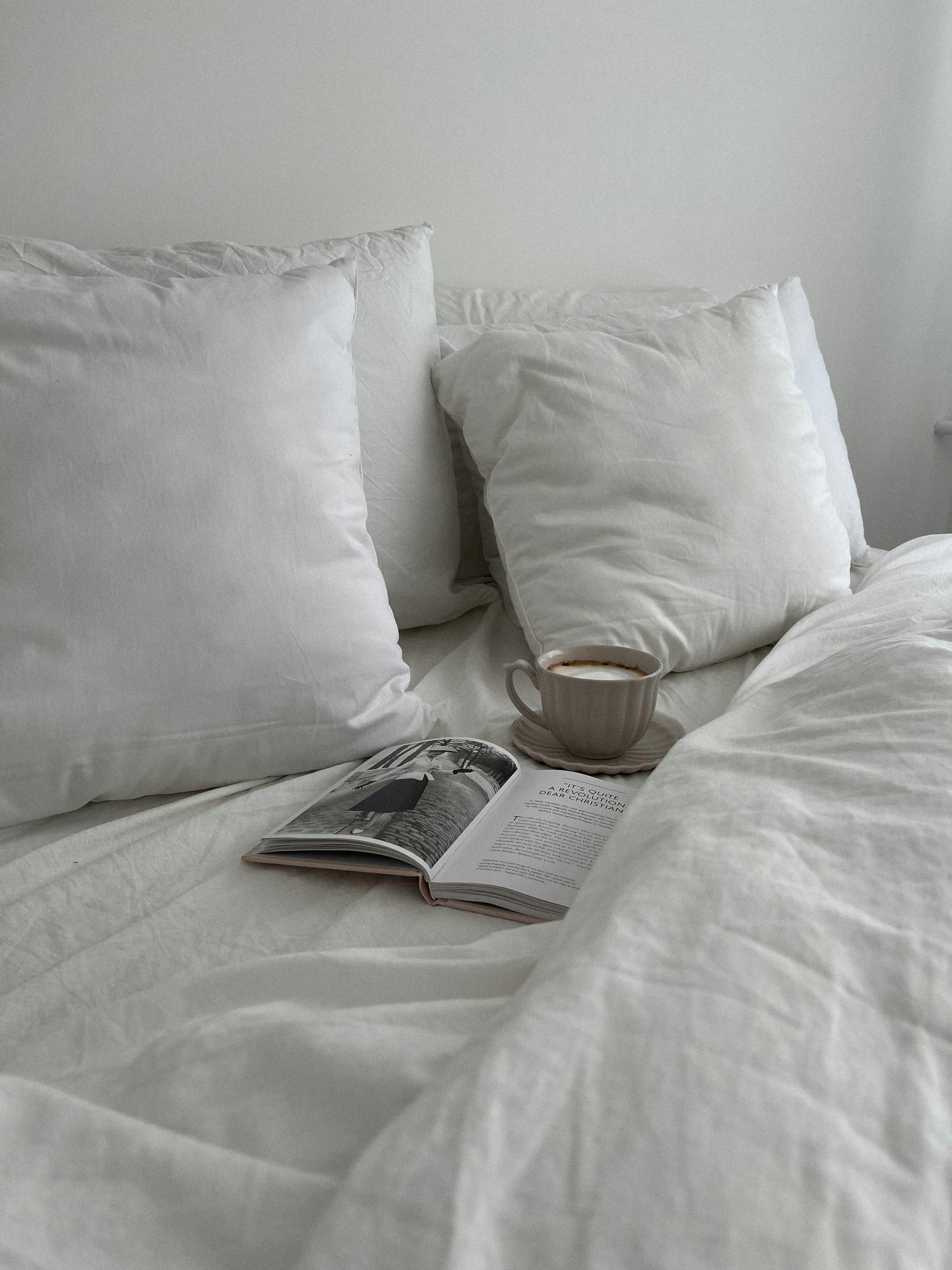 A tranquil bedroom setting with white linens, pillows, a cup of coffee, and an open book on bed.
