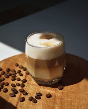 A creamy cappuccino served in a glass on a wooden table with coffee beans scattered around.