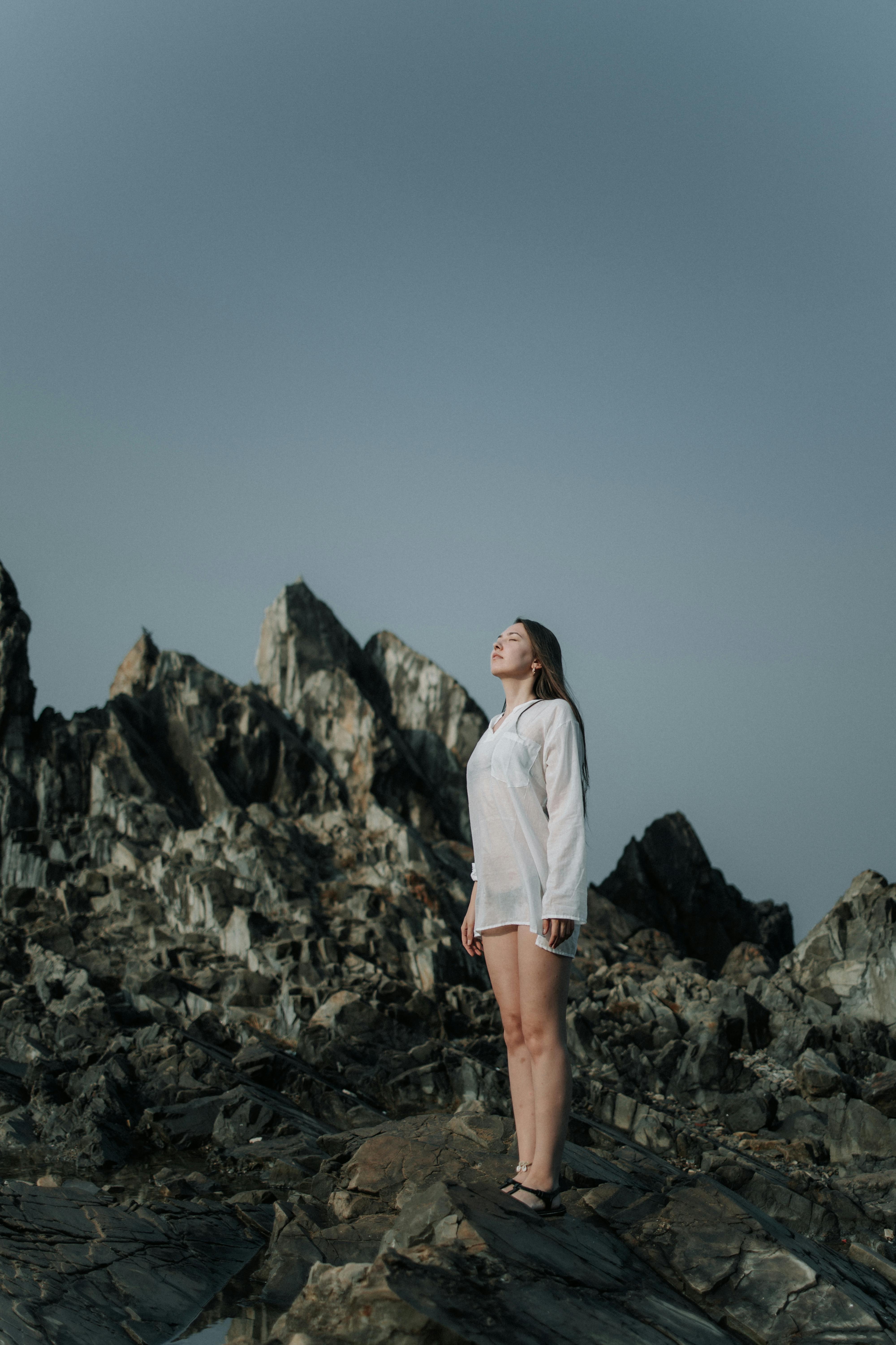 A woman stands confidently on eroded rocks with a serene expression, wearing a white shirt.