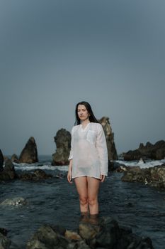 A woman in a white shirt stands in rocky seaside waters under a clear sky.
