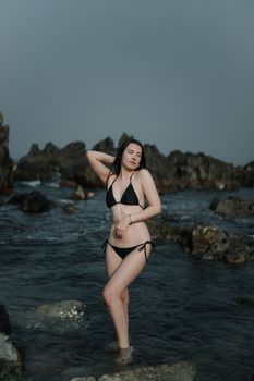 A woman in a black bikini posing by the rocky sea shore during daylight.
