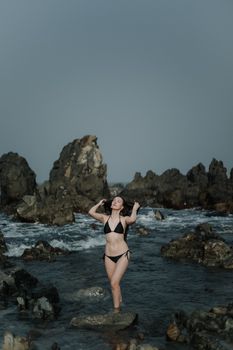 A stylish bikini model posing confidently among scenic rocky shore and sea.