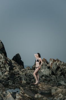A woman in a bikini stands on a rocky shore under a blue sky, embodying summer relaxation.