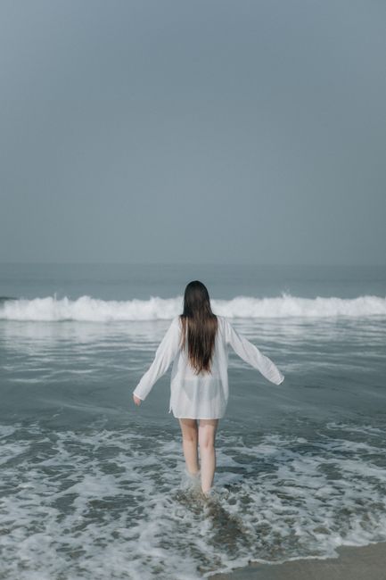A woman with long hair in a white shirt walks into the ocean, capturing a serene and peaceful moment at the beach.