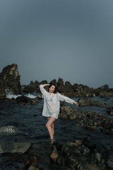 A woman in a white shirt poses artistically on a rocky seashore, capturing a serene and moody atmosphere.