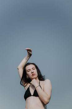 Portrait of a woman with eyes closed, arm raised, standing outdoors in swimwear.