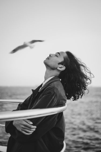 Black and white photo of a man leaning back, eyes closed, enjoying the sea breeze with a seagull in flight.