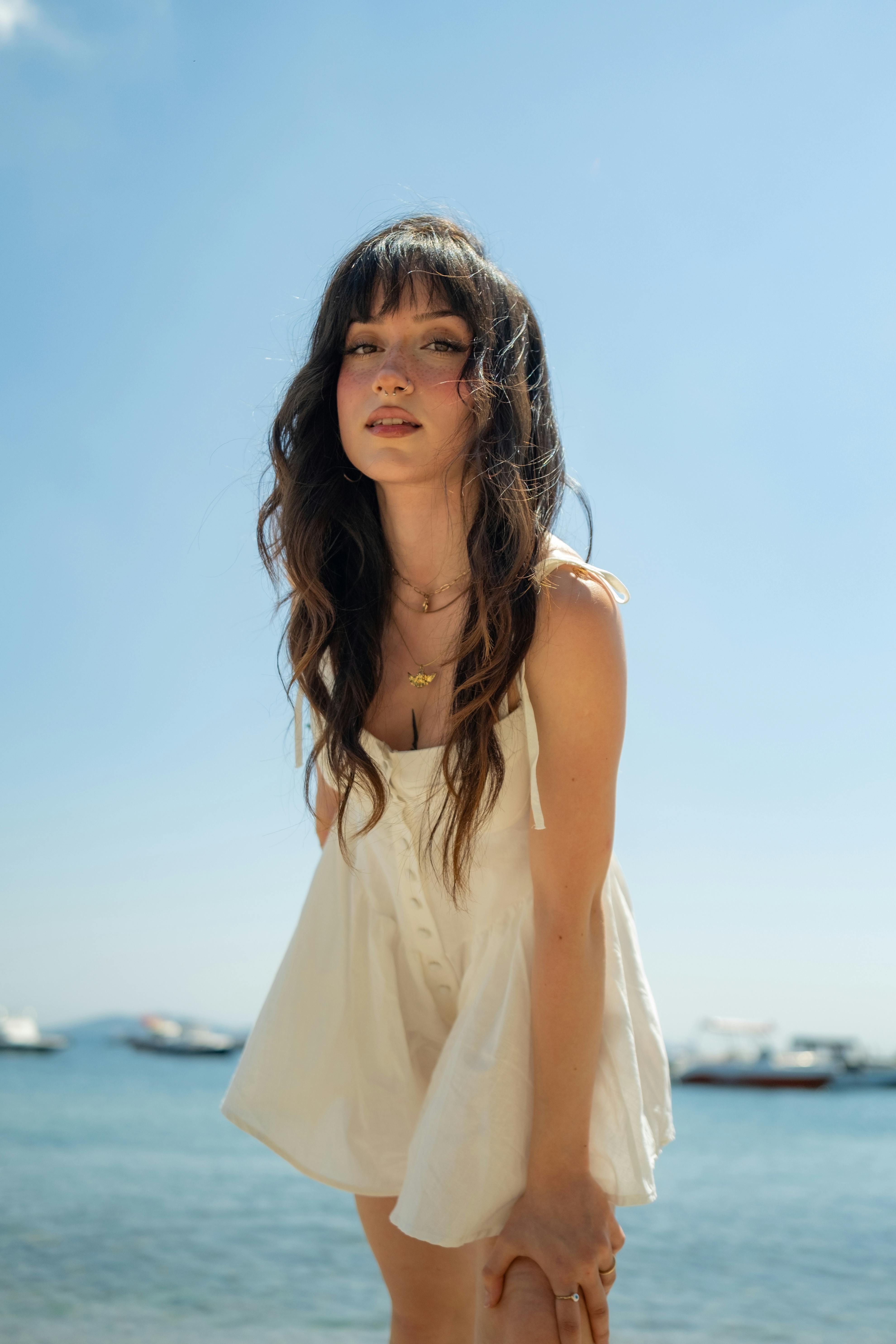 Beautiful brunette standing by the coast in a white dress on a sunny day.