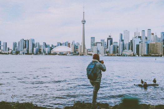 A backpacker taking photos of the Toronto skyline from across the water, featuring the CN Tower.