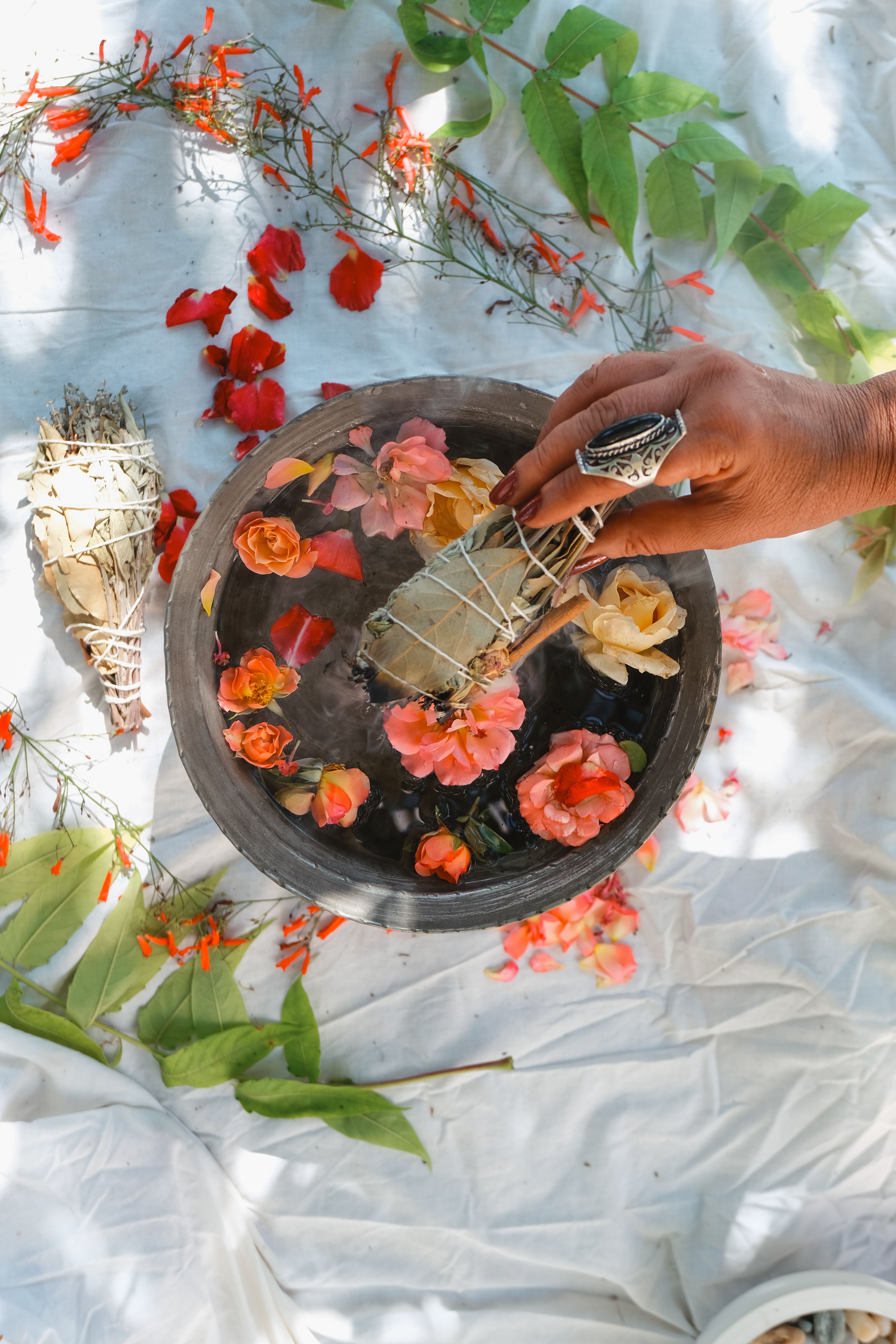 A hand holding a sage bundle over a bowl of water with colorful flowers, embodying tranquility and cleansing.