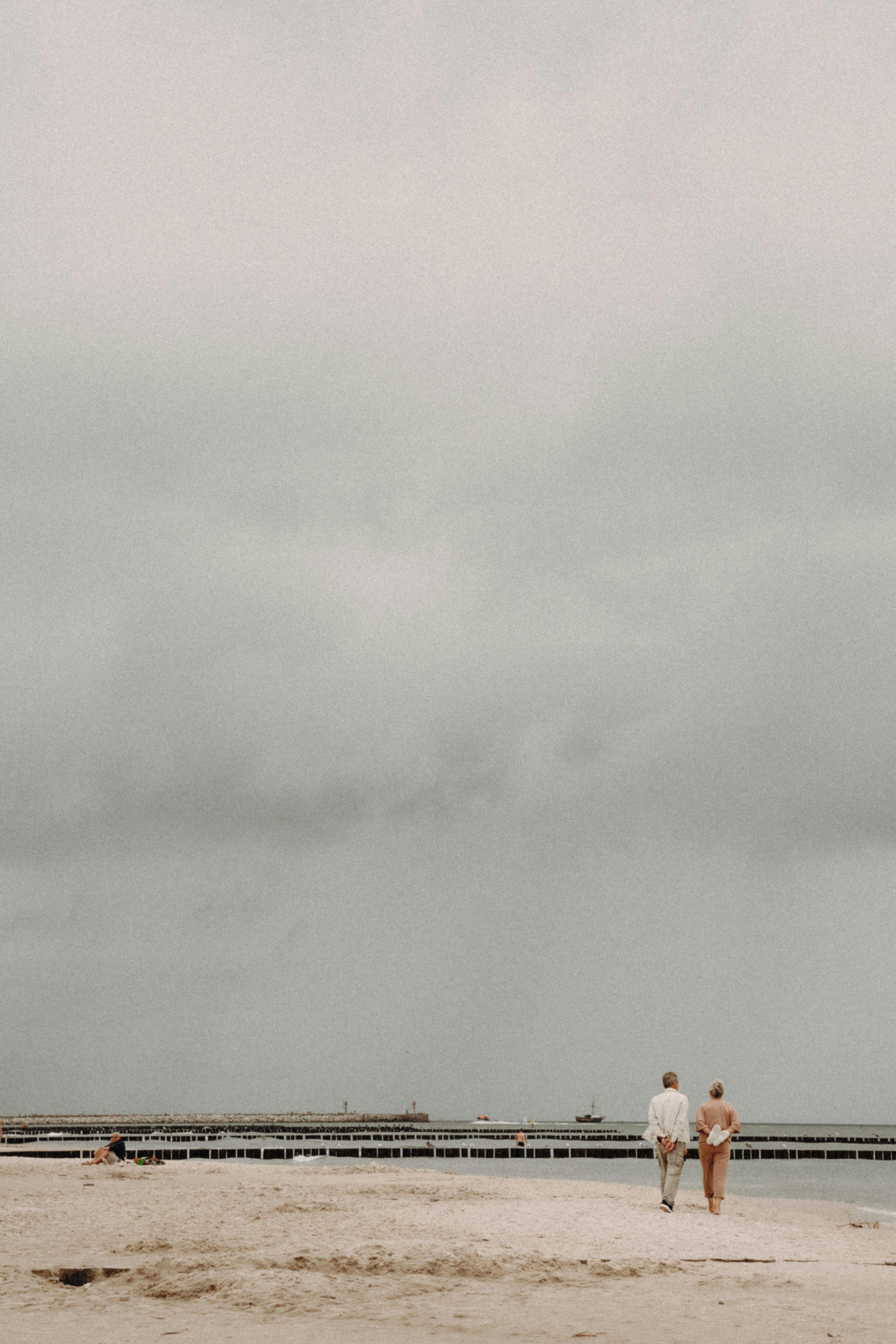 A couple walks along a tranquil beach under an overcast sky, facing the sea.