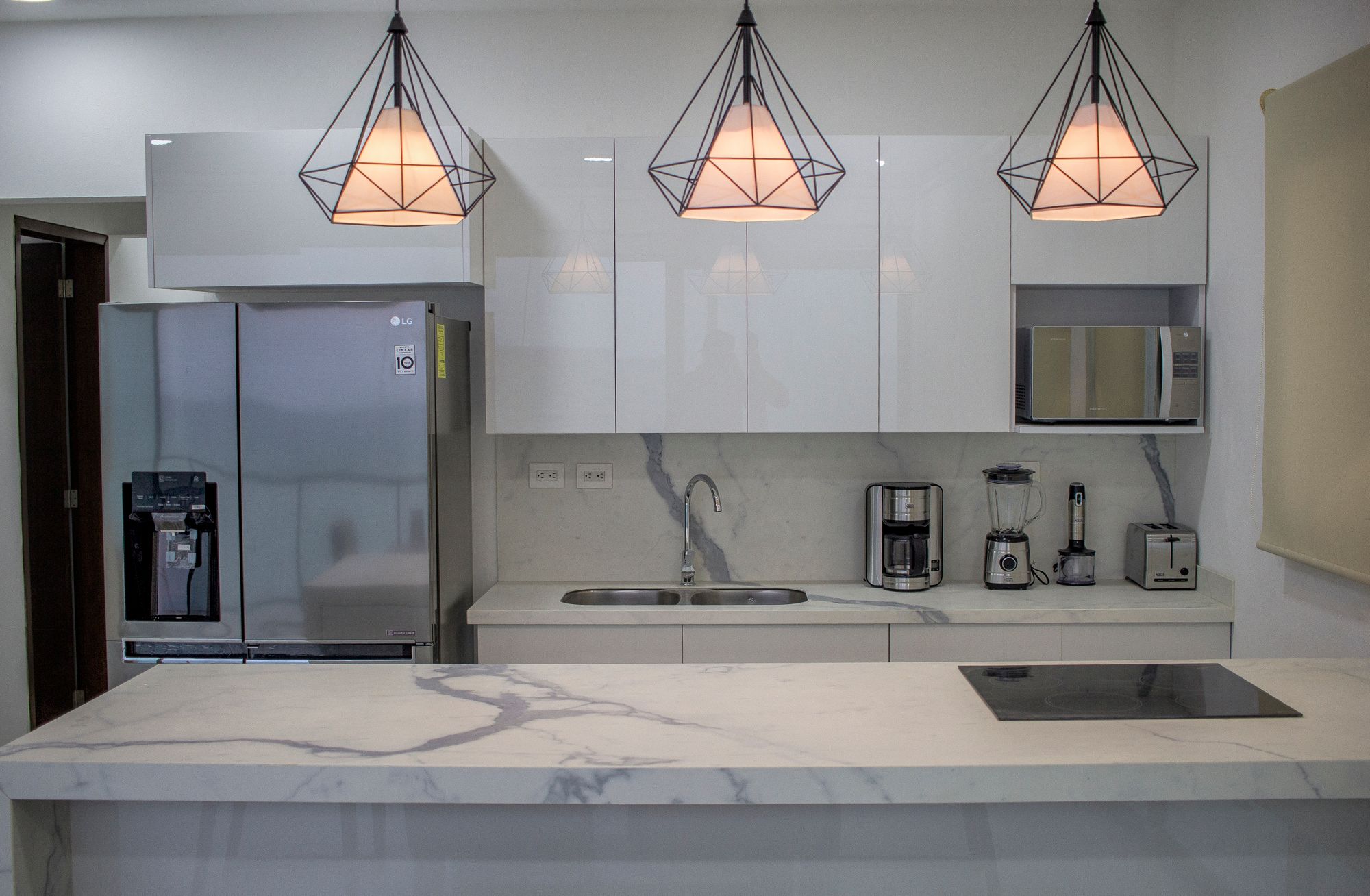 Contemporary white kitchen with marble-look benchtops and splashback, glossy cabinets, stainless-steel refrigerator and microwave, sink, cooktop and pendant lights.