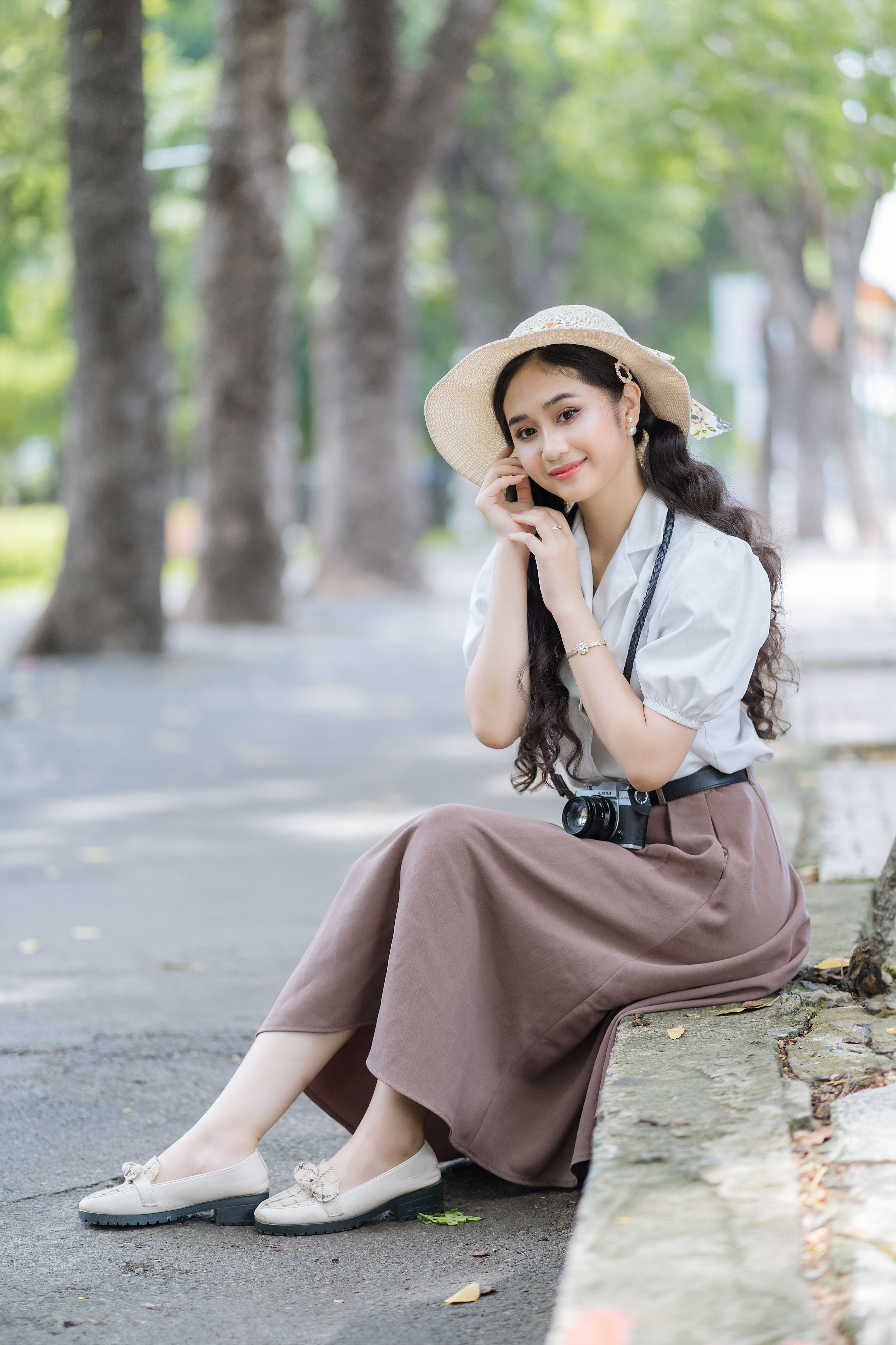Smiling woman in summer attire sitting by the sidewalk with a camera outdoors.