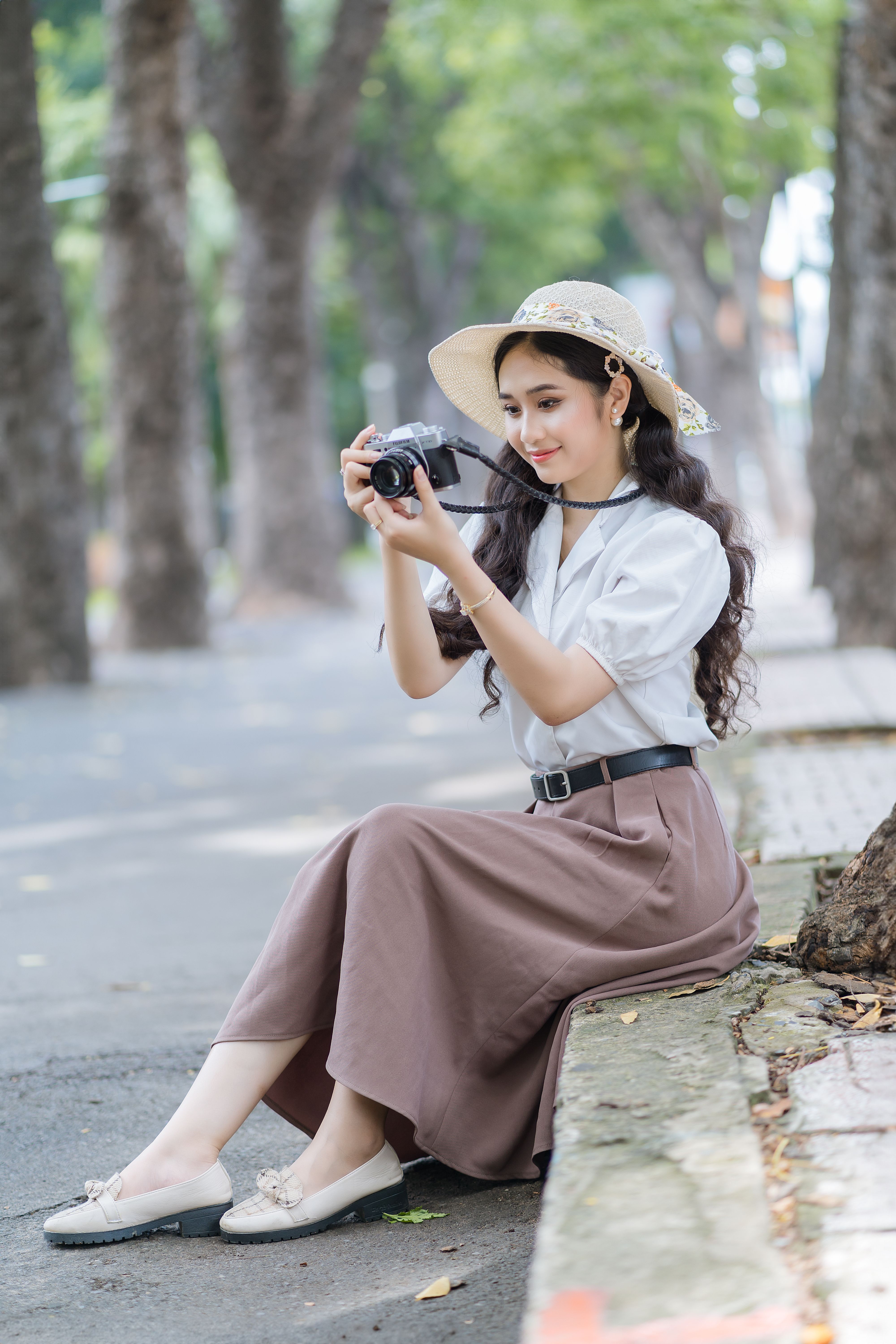 Elegant young woman sitting in a sunny park taking pictures with a camera.