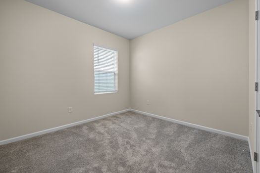 A modern empty room with beige walls, carpet flooring, and one window.