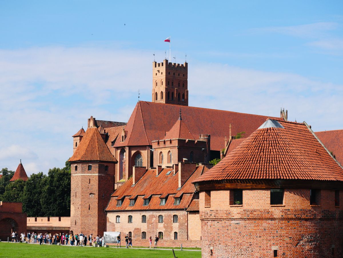 A line of visitors walking the lawn below the castle, whose tall square battlemented tower flies the Polish flag, with a low round brick tower in front.