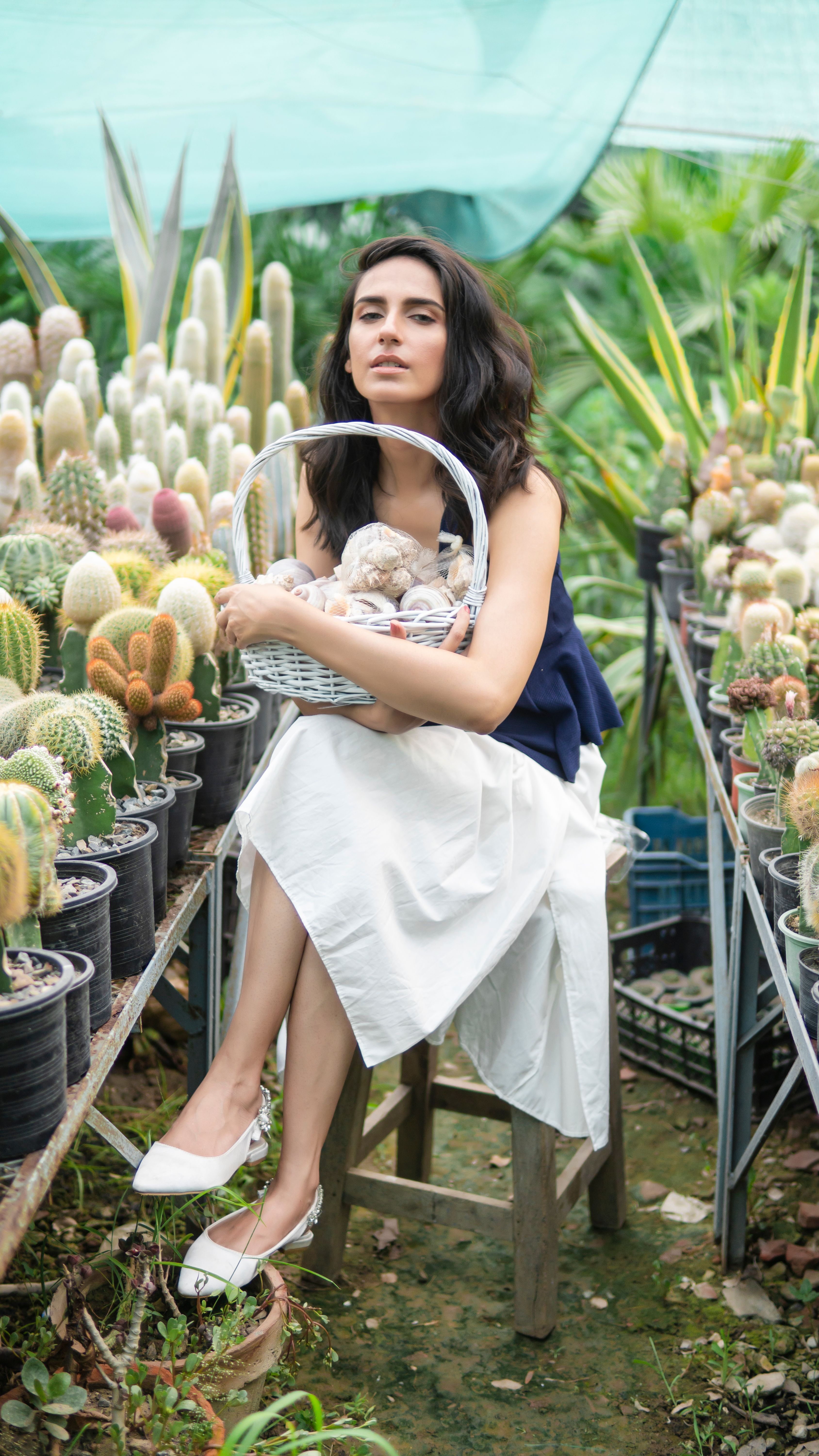 A woman sits among various cacti, holding a basket and showcasing modern fashion in a greenhouse setting.