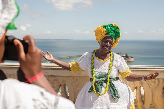A woman in vibrant traditional attire smiles by the sea in Salvador, Brazil, enjoying a sunny day.