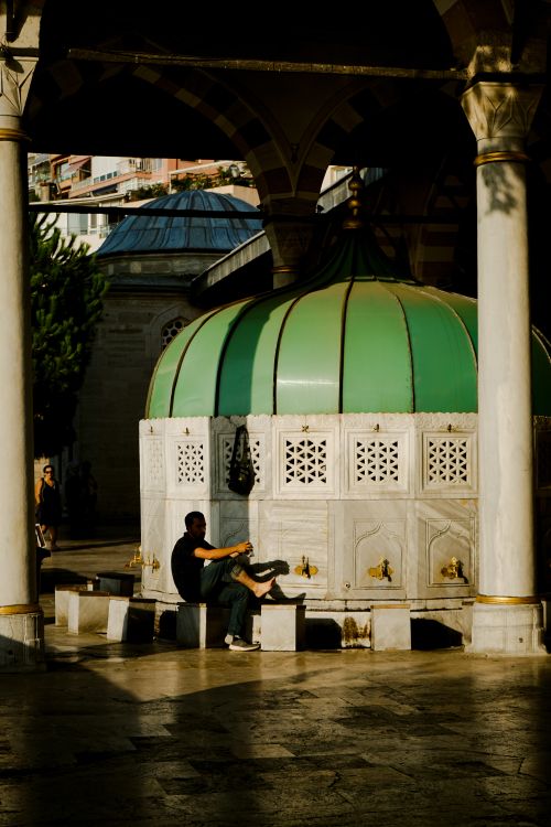 Free A man cleanses his feet at a mosque's ablution fountain under a warm daylight setting. Stock Photo