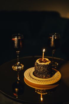 Moody celebration with a lit candle atop a cake and two glasses of red wine in dim lighting.