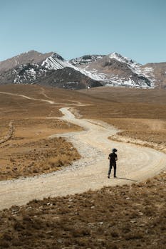 A lone traveler walking on a winding road through a mountainous desert landscape.