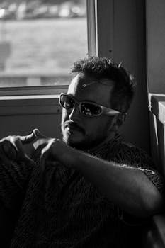 Moody black and white portrait of a man sitting by a train window, wearing sunglasses.