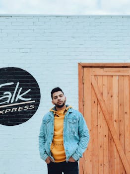 一名时尚男子身穿牛仔夹克和连帽衫，倚靠在砖墙上摆姿势