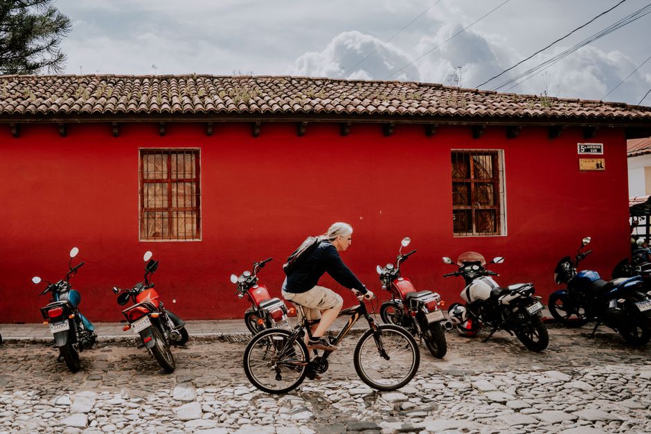 A man rides a bicycle past parked motorcycles on a cobblestone street in front of a red building.