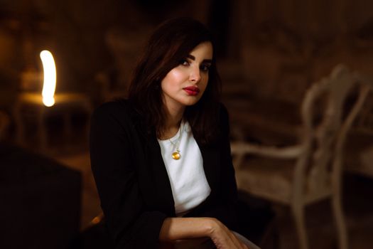 Elegant brunette woman with red lipstick in a dark room, wearing a stylish black jacket and white shirt.