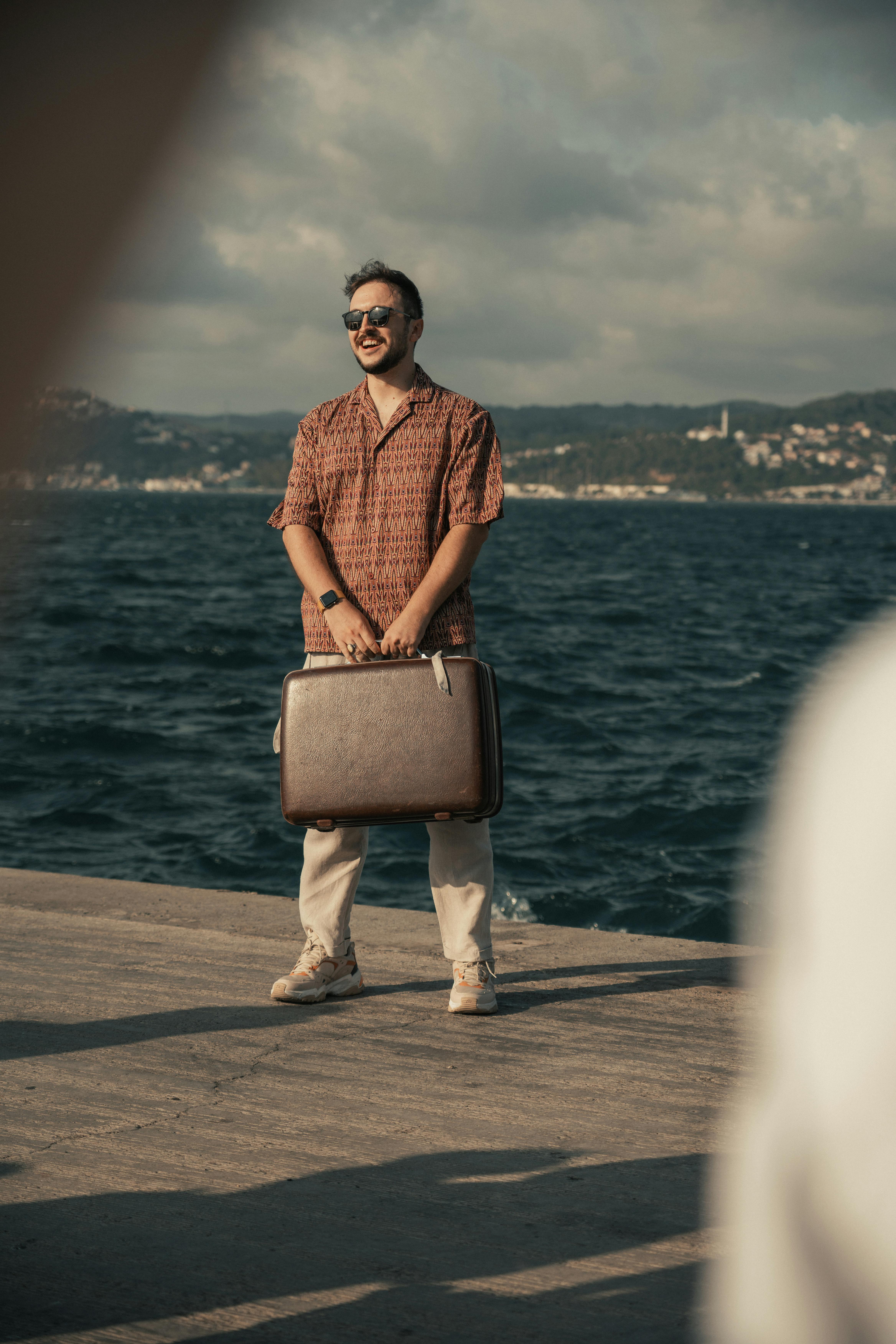 Casually dressed man holding a suitcase on a seaside pier with scenic mountain backdrop.